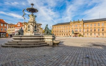 Prunkvoller Barockbrunnen mit Bronzestatuen auf dem Erlanger Schlossplatz. Großes gelbes Schlossgebäude im Hintergrund, Kopfsteinpflaster unter blauem Himmel.