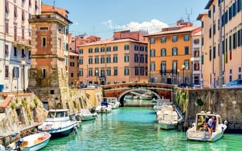Türkisfarbener Kanal in Venezia Nuova, Livorno mit festgemachten Booten entlang Steinmauern. Bunte Gebäude säumen beide Seiten, mit Steinbrücke in der Mitte.