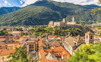 Festung Castelgrande mit Türmen auf einem Felsenhügel über Bellinzonas Altstadt mit roten Ziegeldächern, umgeben von grünen Bergen unter blauem Himmel.