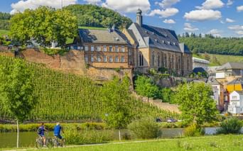 Zwei Radfahrer auf einem Weg neben einem Fluss mit der St. Laurentius Kirche auf einem Hügel in Saarburg, umgeben von Weinbergen und grüner Landschaft.