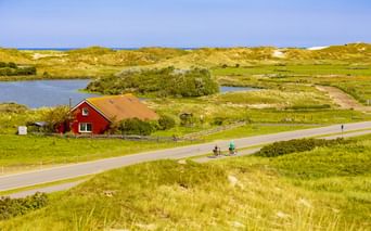 Radfahrer auf asphaltiertem Weg durch Dünenlandschaft auf Norderney mit rotem Haus, Teich und Sanddünen unter blauem Himmel.