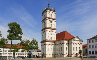 Weiße Stadtkirche mit hohem Turm und rotem Dach am Marktplatz in Neustrelitz, umgeben von historischen Gebäuden und Bäumen unter blauem Himmel.