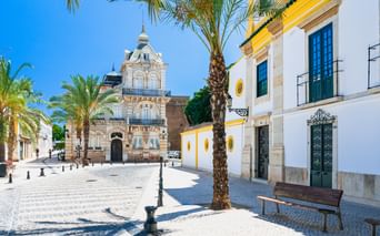 Sonniger Kopfsteinpflasterplatz in Faro mit Palmen, historischer Kirche mit verzierter Fassade und weißen Gebäuden mit gelben Akzenten.
