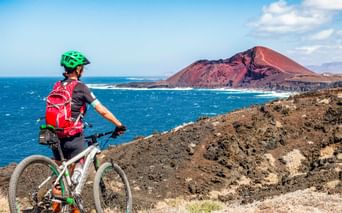 Mountain biker with green helmet and red backpack on rocky terrain overlooking blue ocean and red volcanic cone in Lanzarote.