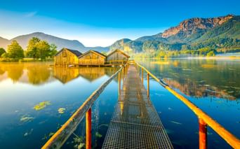 Metallsteg führt zu drei Holzbootshäusern am ruhigen Kochelsee. Berge und Bäume spiegeln sich im stillen Wasser unter blauem Himmel.