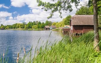 Traditionelles reetgedecktes Bootshaus auf Stelzen an einem ruhigen See mit Schilf im Vordergrund. Blauer Himmel mit Wolken und grüne Bäume säumen das Ufer.