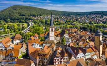 Luftaufnahme von Lohr am Main mit historischem Stadtkern, Kirchturm, roten Ziegeldächern, Fachwerkhäusern und bewaldeten Hügeln im Hintergrund.