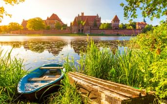 Marienburg in Malbork, Polen, über einen ruhigen Fluss mit blauem Boot und Holzbank im Vordergrund, eingerahmt von grünem Laub.