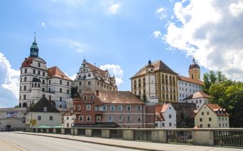 Blick auf die Neuburger Schlossanlage mit weißem Renaissanceschloss, Kirchturm und bunten historischen Gebäuden an der Donau unter blauem Himmel.