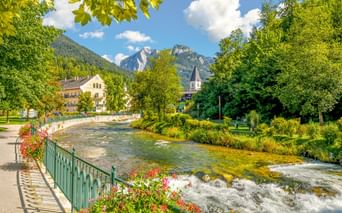 Fluss fließt durch Bad Aussee mit blumengeschmückter Promenade, Kirchturm und Bergen unter blauem Himmel mit weißen Wolken.