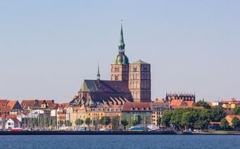 Blick über den Hafen von Stralsund mit bunten historischen Gebäuden am Ufer und der markanten St. Nikolaikirche mit ihrem Kupferturm.