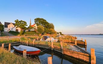 Holzsteg mit vertäuten Booten in Maasholm bei Sonnenuntergang. Kirche mit Turm und Häuser auf dem Hügel, ruhiges Wasser spiegelt den Himmel.
