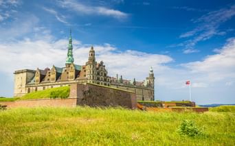 Schloss Kronborg in Helsingør mit grünen Kupfertürmen und Renaissance-Türmen, umgeben von Grashängen. Dänische Flagge weht an hohem Mast.