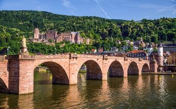 Historische Steinbrücke mit mehreren Bögen über den Neckar in Heidelberg. Ruinen des roten Sandsteinschlosses auf bewaldetem Hang sichtbar.