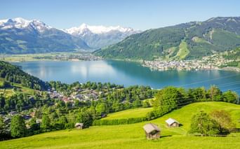 Luftaufnahme des Zeller Sees umgeben von grünen Wiesen mit Holzhütten, bewaldeten Bergen und schneebedeckten Alpengipfeln unter blauem Himmel.