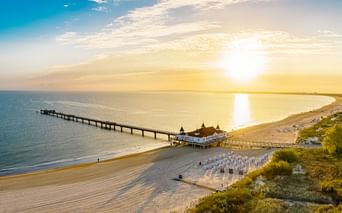 Aerial view of Ahlbeck Pier on Usedom extending into the Baltic Sea at sunset, with sandy beach, beach chairs, and golden sunlight reflecting on water.