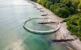 Luftaufnahme eines kreisförmigen Holzstegs, der ins Meer in Aarhus ragt. Die Struktur bildet einen vollständigen Kreis mit Besuchern.