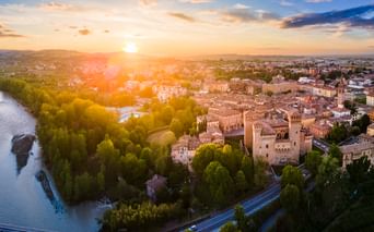 Luftaufnahme der Burg Rocca di Vignola umgeben von Bäumen und historischer Stadt bei Sonnenuntergang. Links fließt ein Fluss durch grüne Landschaft.