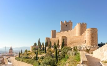 Burg La Atalaya in Villena mit Steinmauern und Türmen auf einem Hügel, umgeben von Zypressen und einem gewundenen Weg unter klarem blauem Himmel.