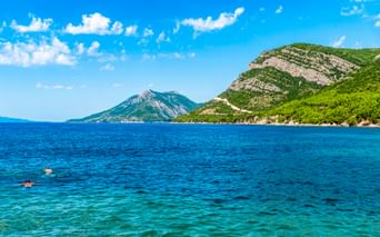 Zwei Schwimmer im klaren türkisfarbenen Wasser nahe der Halbinsel Pelješac. Grüne bewaldete Berge erheben sich von der Küste unter blauem Himmel.
