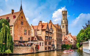 Mittelalterliche Backsteingebäude entlang eines Kanals in Brügge mit dem ikonischen Belfried im Hintergrund. Ein Touristenboot fährt auf dem Wasser.