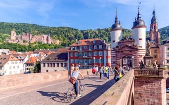Cyclist on Heidelberg's Old Bridge with twin gate towers, historic buildings, and Heidelberg Castle on forested hillside in background.