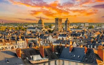 Luftaufnahme von Tours bei Sonnenuntergang mit den Türmen von Saint-Martin-de-Tours über den Dächern. Dramatischer orange-violetter Himmel beleuchtet die Altstadt.
