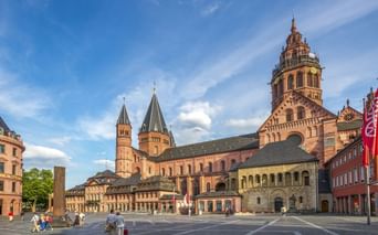 Mainzer Dom mit roten Sandsteintürmen am Domplatz unter blauem Himmel. Historische Gebäude und Fußgänger auf dem Platz.