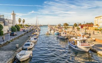 Hafenkanal in Viareggio gesäumt von Fischerbooten und Yachten. Ein weißer Leuchtturm steht links, Palmen und Gebäude auf beiden Seiten unter blauem Himmel.