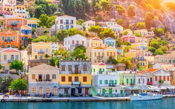 Bunte neoklassizistische Häuser in Gelb, Weiß, Blau und Orange säumen die Uferpromenade der Insel Symi mit einem Boot im Hafen.