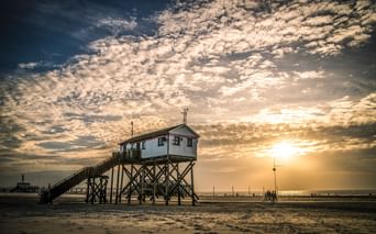 Hölzernes Pfahlhaus mit Treppe am Nordseestrand bei Sonnenuntergang. Dramatischer bewölkter Himmel mit goldenem Sonnenlicht.