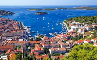 Luftaufnahme des Hafens von Hvar in Kroatien mit rotgedeckten Gebäuden, zahlreichen Booten im blauen Wasser und grünen Inseln in der Ferne.
