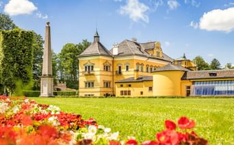 Gelbes Schloss Hellbrunn in Salzburg mit gepflegten Rasenflächen, bunten Blumenbeeten und einem Obelisken unter blauem Himmel mit weißen Wolken.