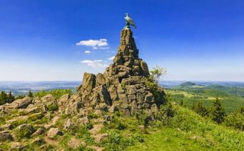Steinpyramide mit Adlerskulptur auf dem Gipfel der Wasserkuppe, mit Blick auf grüne Hügel unter blauem Himmel.