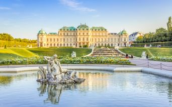 Barockes Schloss Belvedere in Wien mit verziertem Brunnenskulptur im Vordergrund, gepflegten Gärten und türkisfarbenen Kuppeldächern.