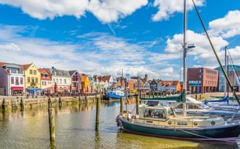 Husumer Hafen mit bunten historischen Häusern entlang der Uferpromenade. Segelboote und Yachten liegen im Hafen unter blauem Himmel mit weißen Wolken.