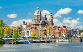 Amsterdamer Waterfront mit den Türmen der St. Nicolaaskerk über bunten Grachtenhäusern. Hausboote säumen den Hafen unter blauem Himmel.