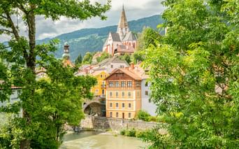 View of Murau old town in Austria with a prominent church spire, colorful historic buildings along a river, framed by green trees and mountains.