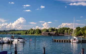 Hafen am Bodensee mit Booten und Holzstegen. Historische Pfahlbauten des Pfahlbaumuseums sind über das Wasser hinweg unter blauem Himmel sichtbar.