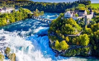 Luftaufnahme des Rheinfalls bei Schaffhausen mit tossendem Wasser, historischem Schloss, Steinbrücke und grünen Wäldern unter blauem Himmel.