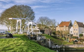Weiße hölzerne Zugbrücke mit traditionellen niederländischen Häusern entlang eines Kanals in Schoonhoven. Grüne Grasuferböschung und kahle Bäume unter blauem Himmel.