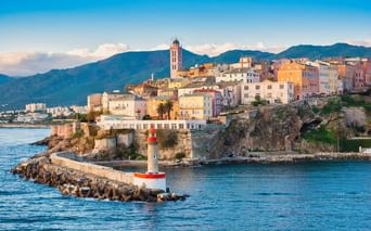 Rot-weißer Leuchtturm auf Steinmole im Hafen von Bastia mit bunten Altstadthäusern auf Klippe und Bergen im Hintergrund.