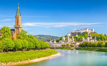 Salzburger Stadtbild mit Salzach, grünen baumgesäumten Ufern, Kirchturm und Festung Hohensalzburg auf dem Hügel unter blauem Himmel.