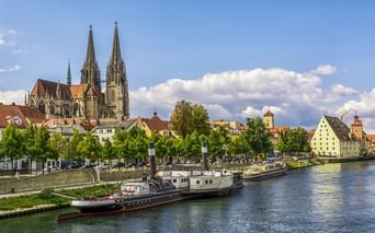 Regensburg Cathedral with twin Gothic spires overlooking the Danube River. Historic boats moored along the waterfront with colorful buildings and trees.