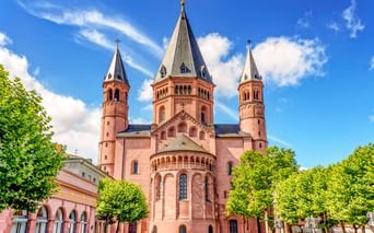 Mainz Cathedral with three red sandstone towers topped with crosses, flanked by green trees under a blue sky with white clouds.