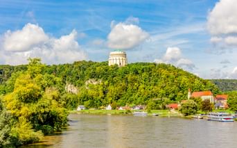The Befreiungshalle monument sits atop a forested hill above Kelheim. The Danube River flows below with boats moored along the green riverbank.
