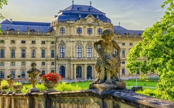 Würzburger Residenz mit prunkvoller Barockfassade und blauem Dach. Steinbrunnen mit Skulptur im Vordergrund, umgeben von grünem Rasen und Bäumen.