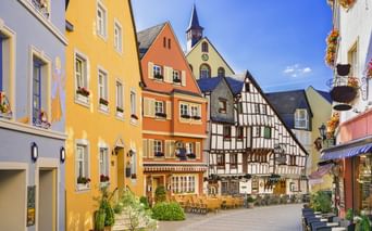 Bunte Fachwerkhäuser und bemalte Fassaden umgeben einen sonnigen Marktplatz in Bernkastel-Kues mit Außengastronomie und Kirchturm.