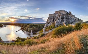 Ruinen der Burg Devin auf einem Felsen mit Blick auf die Donau bei Sonnenuntergang, mit goldenem Gras im Vordergrund und bewaldeten Ufern.