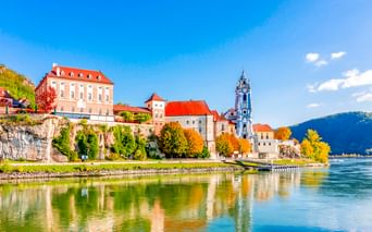 Dürnstein in Wachau with historic buildings and distinctive blue church tower reflected in the Danube River, mountains in background.